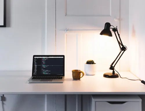 A white desk with an Apple laptop, coffee mug and a lamp.